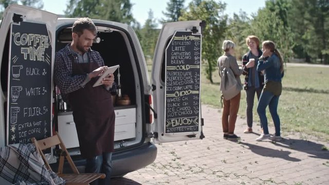 Young male barista with beard wearing apron standing before his mobile coffee van in park and looking at his tablet, group of chatting friends with drinks in background