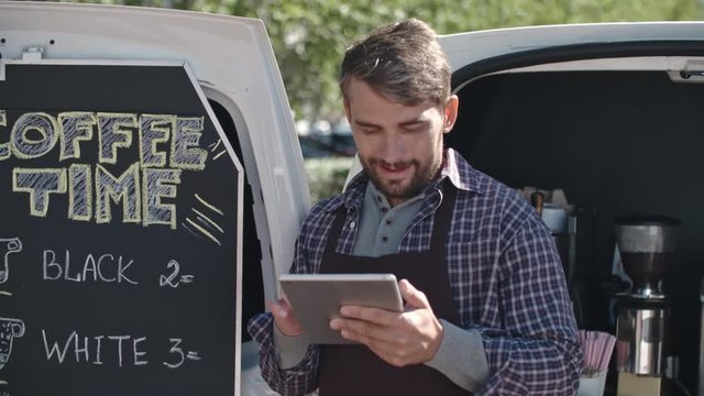 Medium shot of smiling young male barista with beard leaning on doors of his mobile coffee van and browsing social media on tablet