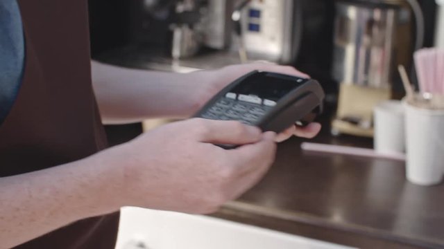 Mid-section of male barista holding portable ATM machine and accepting payment for coffee from female customer touching her mobile phone to its screen 