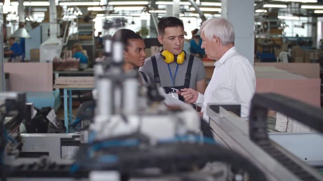 PAN of young Latin-American factory worker standing beside Asian male colleague and discussing work with elderly manager