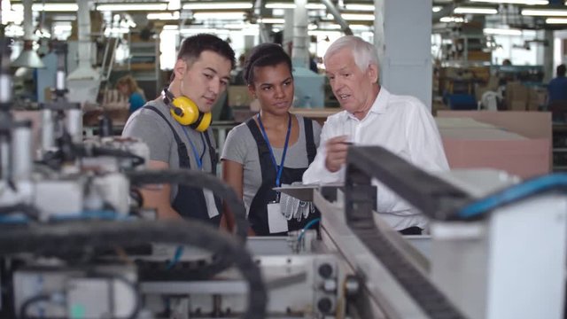 PAN of young Latin-American female worker discussing machinery with Asian colleague and elderly factory supervisor 