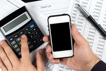 Hands of businessman holding white smartphone and calculating revenue or expenses.