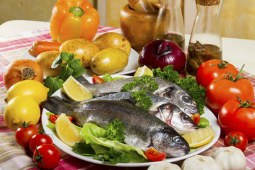 Three pieces of raw and fresh fish, sea bream, sea bass and trout on a white plate beside fresh vegetables and lemon.