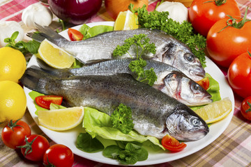 Three pieces of raw and fresh fish, sea bream, sea bass and trout on a white plate beside fresh tomatoes and lemon.