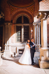 Beautiful sensual brunette bride and handsome groom at sunset castle balcony. Newlyweds near columns and old door embracing and posing kissing picture.