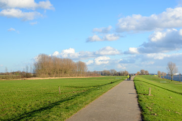 Fototapeta na wymiar Bike and walking path over an embankment along a wide river in the Netherlands. It's a sunny day in the winter season and in the background an unknown elderly couple is strolling together.