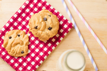 Chocolate chips and chest nut cookies on red and white checked cloth with milk and paper party straws on wood table, top view