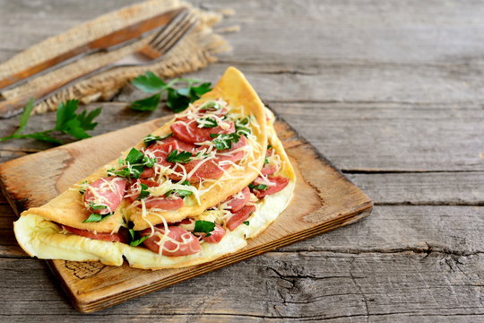 Delicious sausage and cheese omelet on a cutting board and an old wooden background with blank copy space for text. Burlap, cutlery, leaves of fresh parsley. Country breakfast concept
