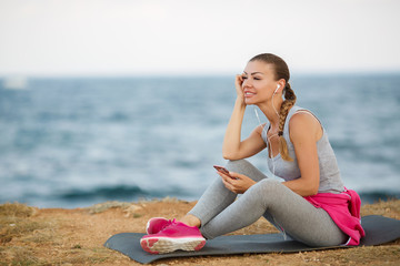 Fototapeta na wymiar Athletic woman,brunette,hair in a braid,dressed in a grey shirt and grey sweat pants,pink sneakers,sitting on fitness Mat in the background of the sea,listening to music through white headphones