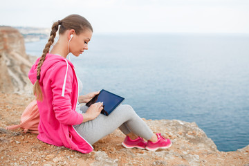 Beautiful woman with long hair in a braid,dressed in grey sweat pants,pink sneakers and a pink sports jacket ,sitting on a rock on a background of blue sea with a tablet in hands