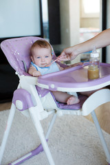 Cute baby with blond hair and blue eyes, wearing a blue shirt, sitting at a table purple for feeding babies, eating applesauce from pink spoon that feeds his mom
