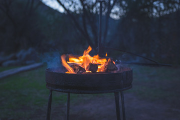 Burning camp fire at dusk in camping site, preparing for barbeque or braai, outdoors activity in South Africa. Selective focus on fire and firewood, very shallow depth of field, cold toned image. - obrazy, fototapety, plakaty