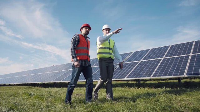 Two male electrician workers in reflective vests walking in between long rows of photovoltaic solar panels