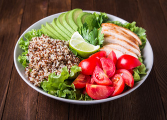 Healthy salad bowl with quinoa, tomatoes, chicken, avocado, lime and mixed greens (lettuce, parsley) on wooden background close up. Food and health.
