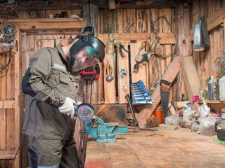 Adult man in work clothes, protective mask and gloves standing near wooden table with a vise and cutting machine saw off a piece of iron on the background of sparks inside the repair shop with tools - obrazy, fototapety, plakaty
