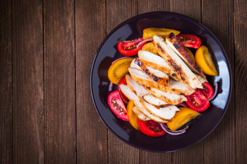 Sliced roasted chicken breast and fresh salad with tomato and purple onion top view on dark wood background. Healthy food. Space for text.