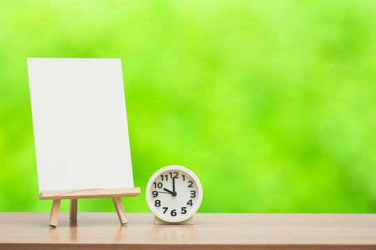 wooden easel with blank canvas and round clock on green nature background