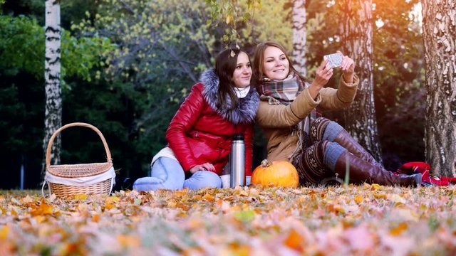 Beautiful brunette girls making selfie on a picnic in autumn park sitting the fallen leaves near the pumpkin at halloween time. 3840x2160