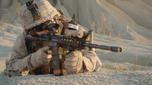 Soldier Lies Down on the Hill, Aims through the Assault Rifle Scope in Desert Environment. Slow Motion.