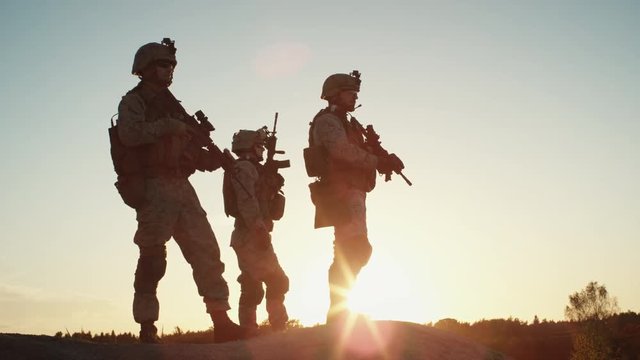 Squad of Three Fully Equipped and Armed Soldiers Standing on Hill in Desert Environment in Sunset Light. Slow Motion.