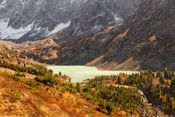 Panoramic view from above on the smooth surface of the turquoise mountain lake, which is located at the foot of the stone wall. Water flowing out of the lake falls down into small river.
