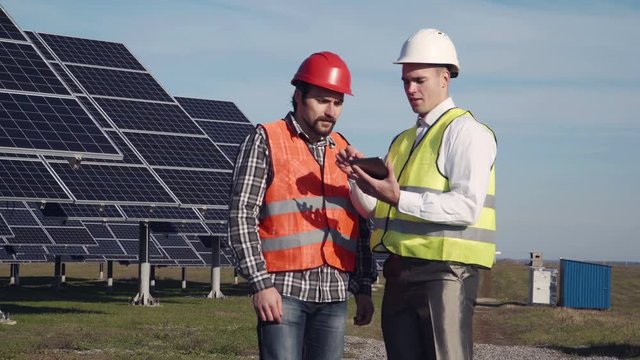 Two engineers in uniform and helmets talking at solar power station and using tablet.
