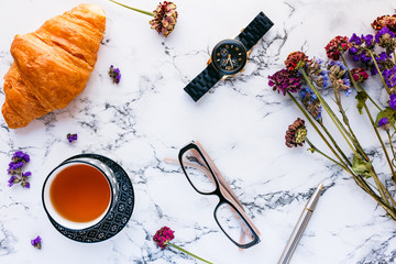 Feminine workspace. Table witn croissant, tea, glasses, watch and flowers on marble background. Lay flat, top view.