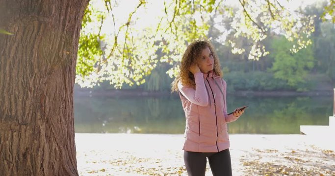 Woman using headphones smart phone listen to music player outdoor sunrise, curly hair sport girl morning autumn park near tree natural sun lights slow motion 60
