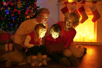 Elderly couple with their granddaughters opening Christmas present near fireplace