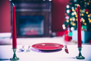 Beautifully laid new year table with red plates and Cutlery on a background of decorated room with Christmas tree and fireplace