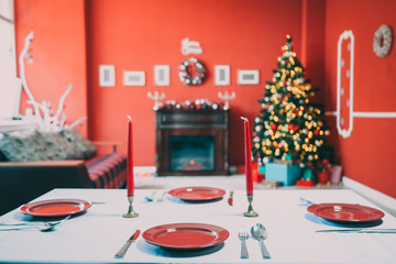 Beautifully laid new year table with red plates and Cutlery on a background of decorated room with Christmas tree and fireplace