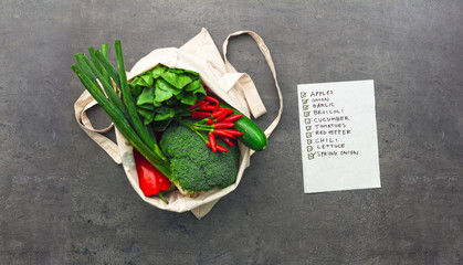 Cotton shopping bag full of vegetables and fruits with checked shopping list. Flat lay food on table.