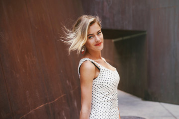 A waist up photo of a charming caucasian blonde woman with hair waving in the wind wearing white dress with black polka dots and smiling at the camera while standing on a brown walls background.