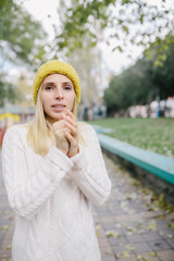 Portrait of freezing young woman in a white woolen sweater and yellow knitted hat standing outdoor in park. woman feels freeze rubs and warms her hands.