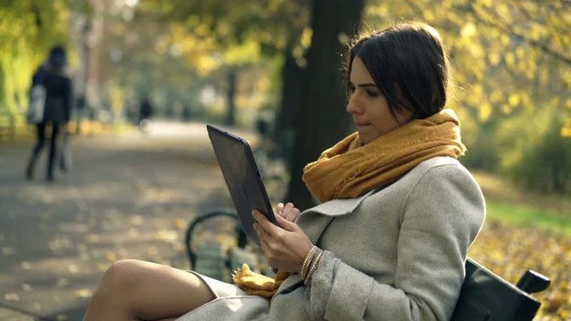 Young woman using tablet computer while sitting on bench in autumn park

