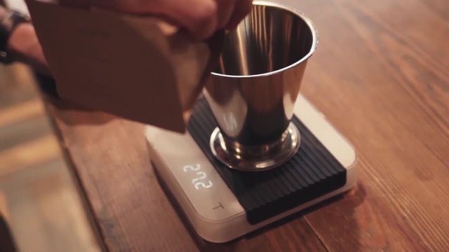 Cafe activities, weighing of roasted coffee beans on the kitchen electronic scales. Barista pours and weights roasted coffee beans on the kitchen electronic scales, on a large oak table.