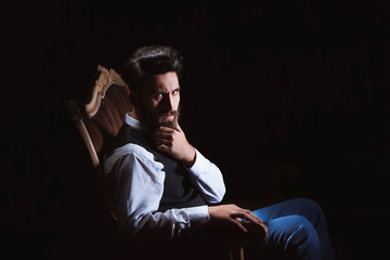 Young handsome bearded caucasian man sitting on vintage sofa. Perfect skin and hairstyle. Wearing vest, white shirt, jeans. Studio portrait with dramatic light.