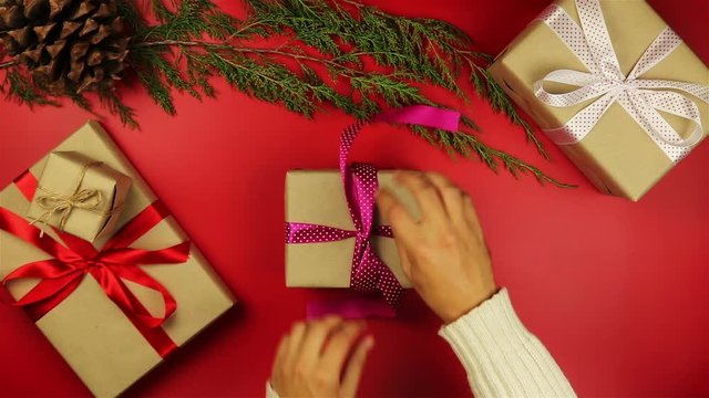 Top view hands unwrapping christmas presents on red background from above. Gift wrapped in brown paper and tied purple ribbon