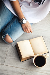 Young modern girl in torn jeans reading a book with a big cup of