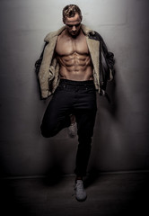 Attractive muscular athlete young man wearing pants and leather jacket looking down while leaning on wall isolated on light gray background
