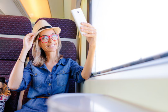 Enjoying travel. Young pretty woman traveling by the train sitting near the window using smartphone, taking selfie.