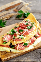 Egg omelette with grated cheese and fried sausages on a board, fork, knife, burlap, fresh parsley leaves on old wooden table. Delicious omelette recipe. Closeup