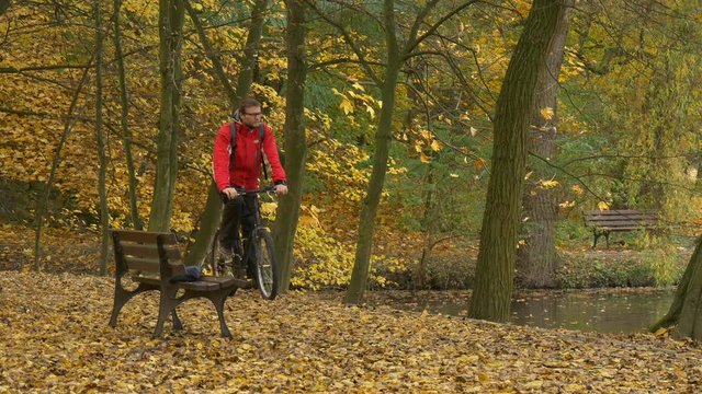 Man Riding a Bicycle Gets Off Sits Down to Bench Autumn Day Cyclist is Resting Among Golden Trees in Park by Lake Surfase is Covered With Yellow Leaves