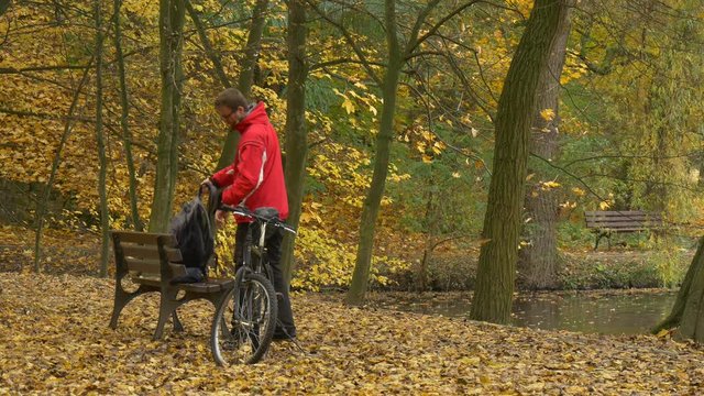 Cyclist Comes Left His Bike Sits Down to Bench Autumn Day Man is Resting Among Golden Trees in Park Ground is Covering With Yellow Leaves Footpath Alley