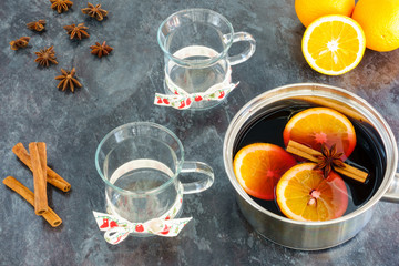 Stainless steel pot with hot, freshly prepared, homemade mulled wine, two mulled wine glasses, cinnamon sticks, star anise and oranges on black marbled kitchen worktop.

