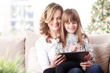 Mother and daughter at christmas, Portrait of cute little daughter using digital tablet while sitting together with her mother on couch at home at christmas time. 