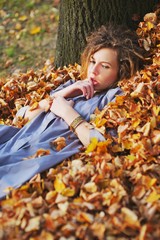 Cute teen girl in a light purple cloak with sad eyes lies on a pile of fallen autumn leaves in the forest, leaning his head against the tree, close up.