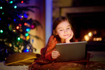 Fototapeta na wymiar Adorable little girl using a tablet pc by a fireplace on Christmas evening