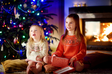 Happy little sisters opening magical Christmas gift by a fireplace