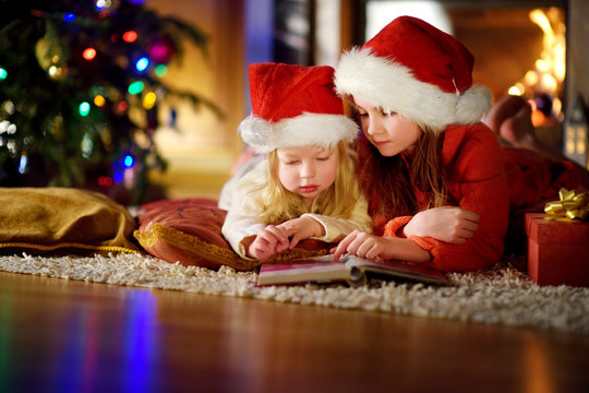 Two cute little sisters reading a story book together under a Christmas tree